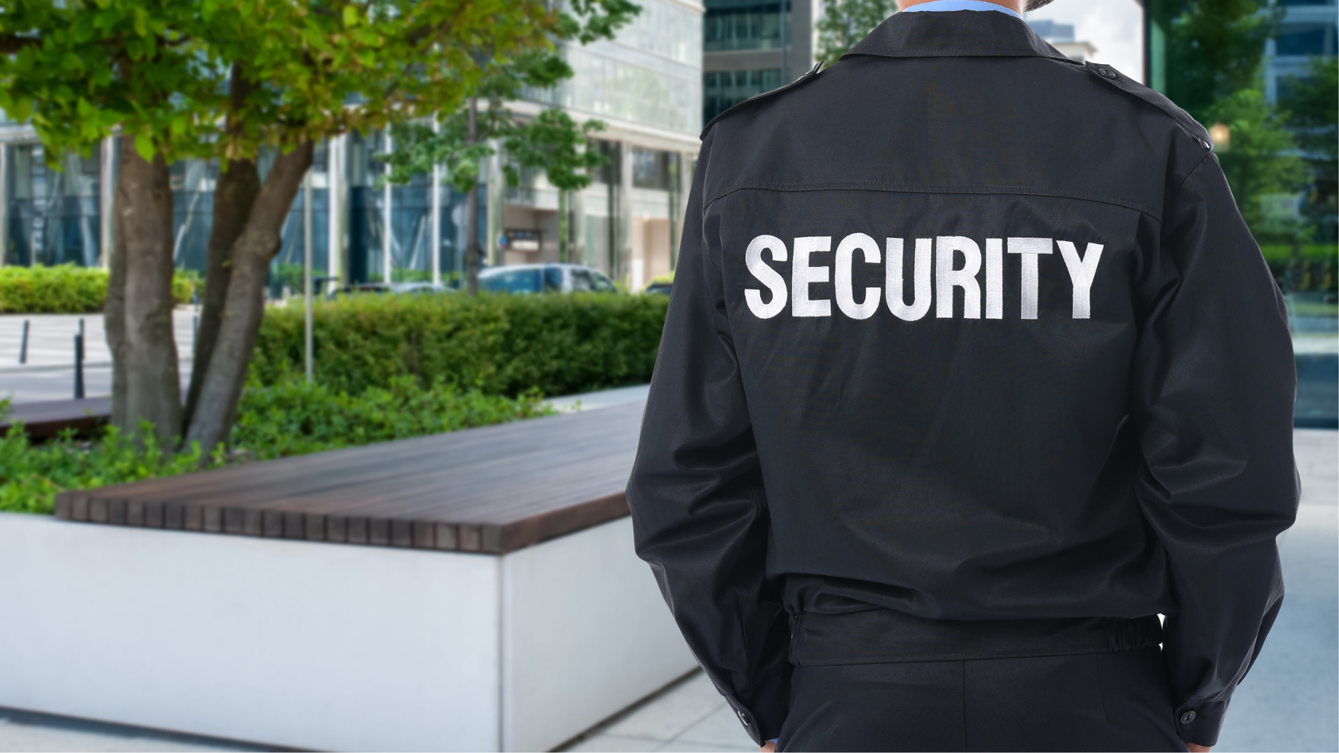 A person in a black security uniform standing outdoors with a green tree and a modern building in the background.