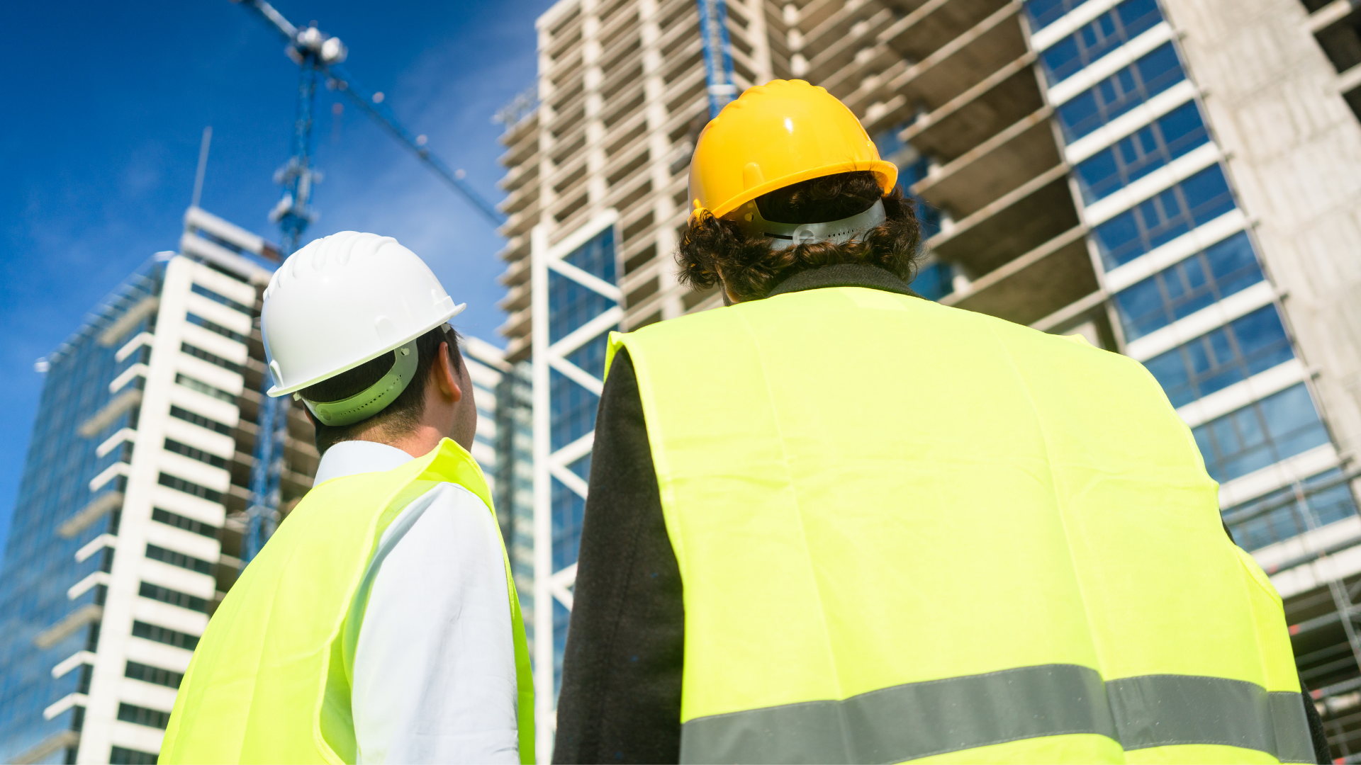 Two construction workers in high-visibility vests and hard hats look up at a large building under construction.