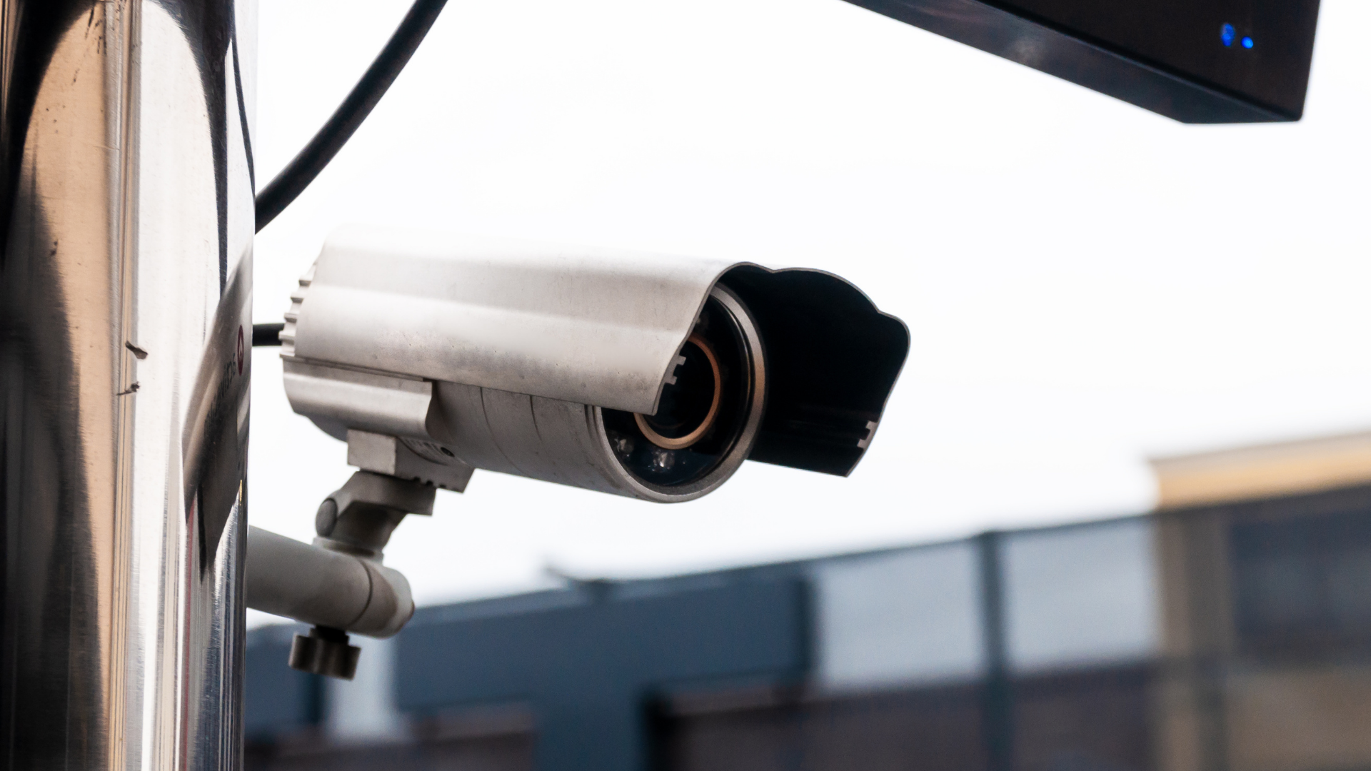 A silver outdoor security camera mounted on a metal pole against a blurred background.