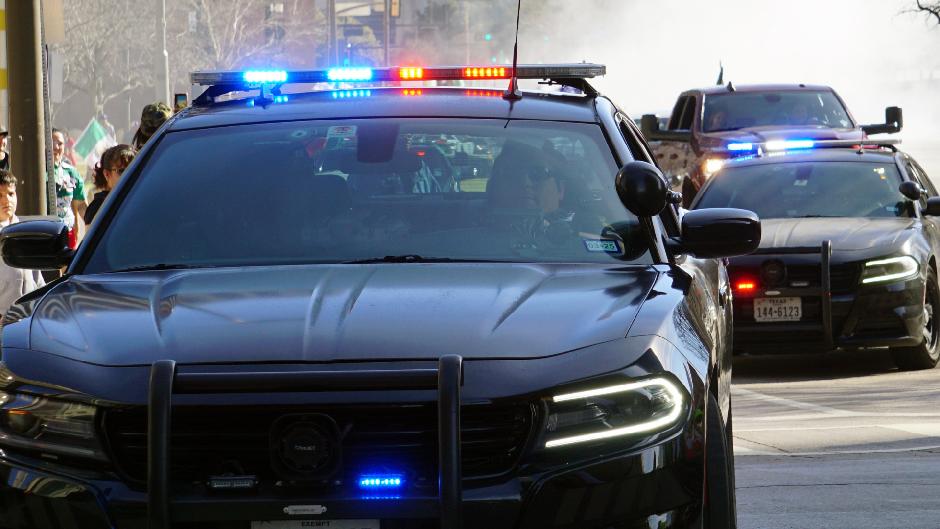 Two black police cruisers with activated blue and red emergency lights traveling in a line on a street.