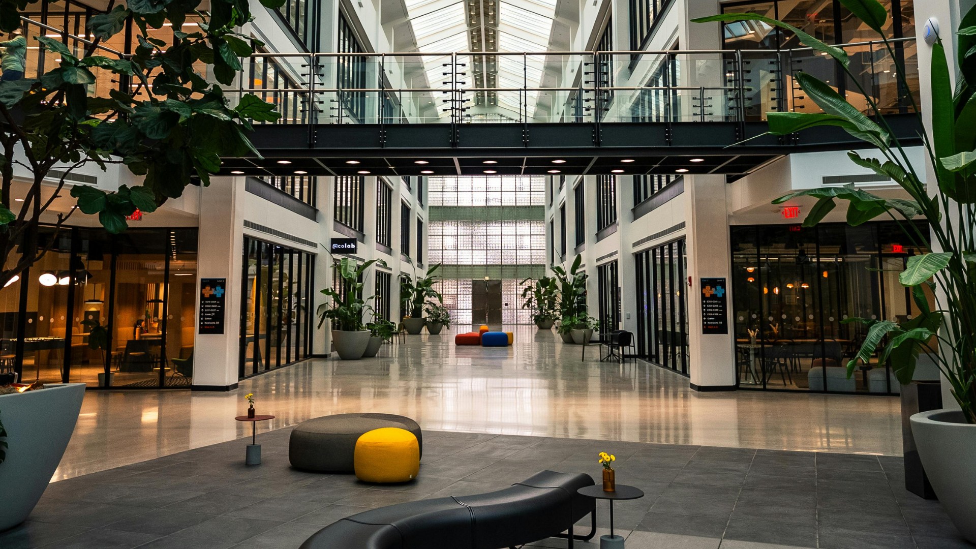 A brightly lit indoor office atrium with high ceilings, glass-walled offices, potted plants, and modern lounge seating.