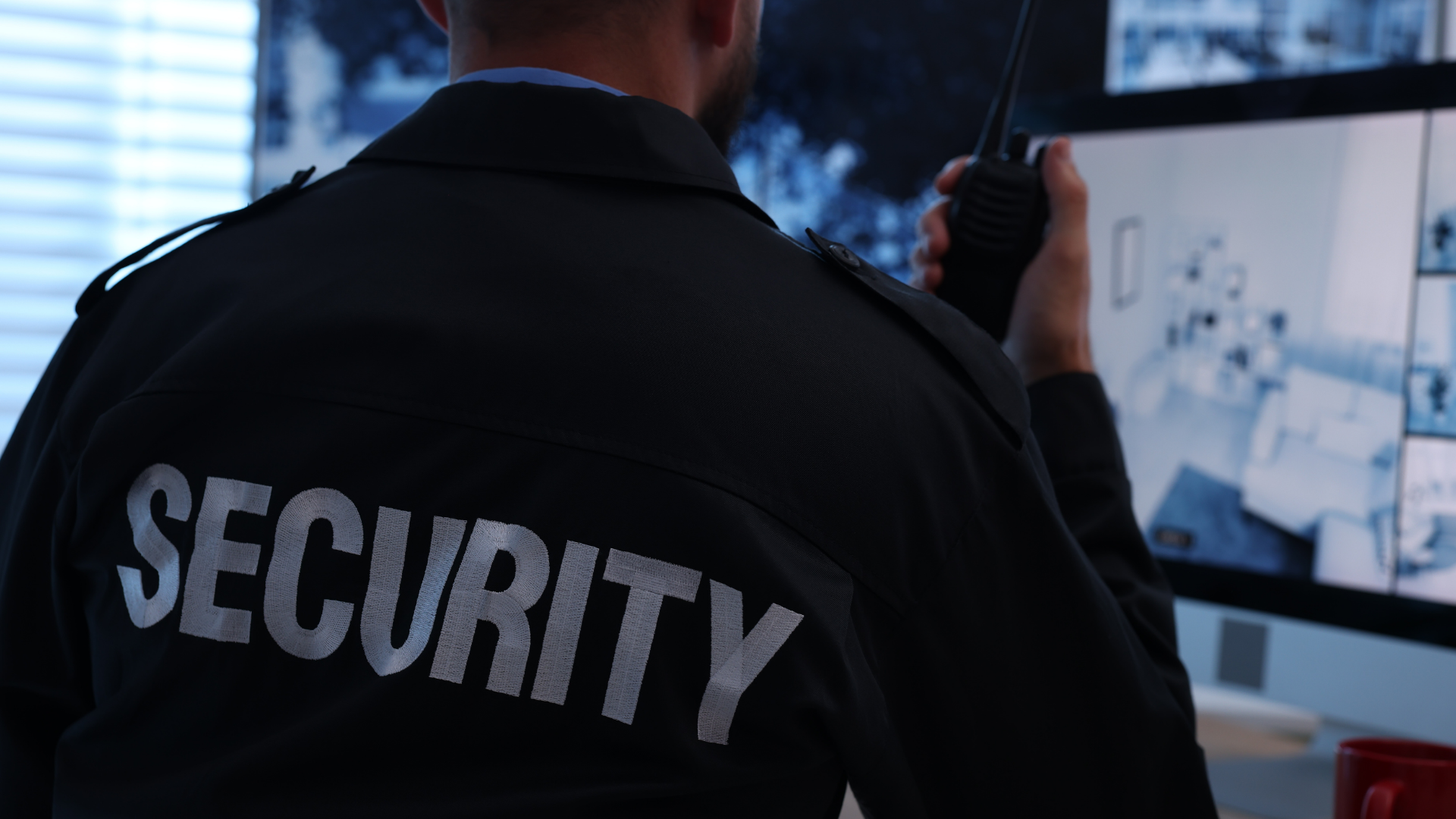 A security guard in a black uniform seen from behind, holding a walkie-talkie while monitoring CCTV screens.