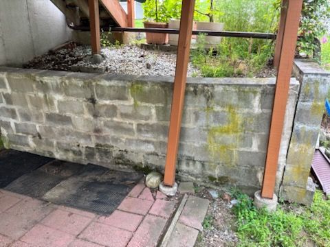 Gray cinder block retaining wall with brown wooden supports, beneath a wooden structure, green vegetation.
