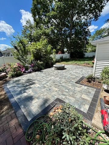 Brick patio with a fire pit, surrounded by gardens and landscaping, on a sunny day.