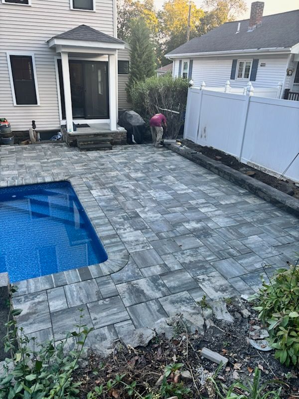 Poolside patio with blue water and workers laying pavers. White fence, house in background.