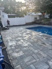 Poolside patio with gray pavers, pool, white fence, and a worker in the background.