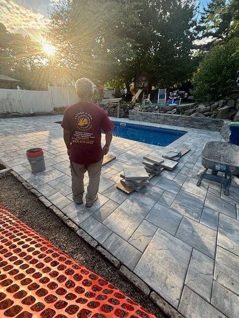 Man in red shirt stands by a pool with new stone patio. Sunshine illuminates the scene.