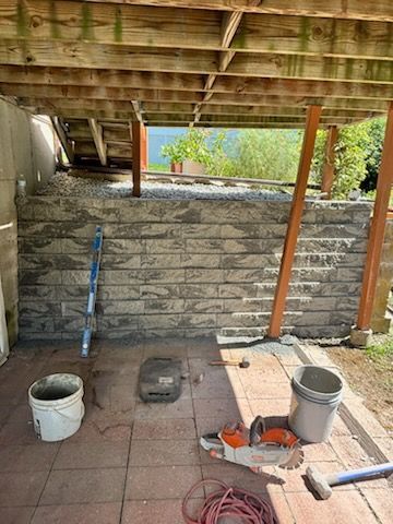 A partially built stone wall under a wooden deck; tools, buckets, and a saw sit on red pavers.