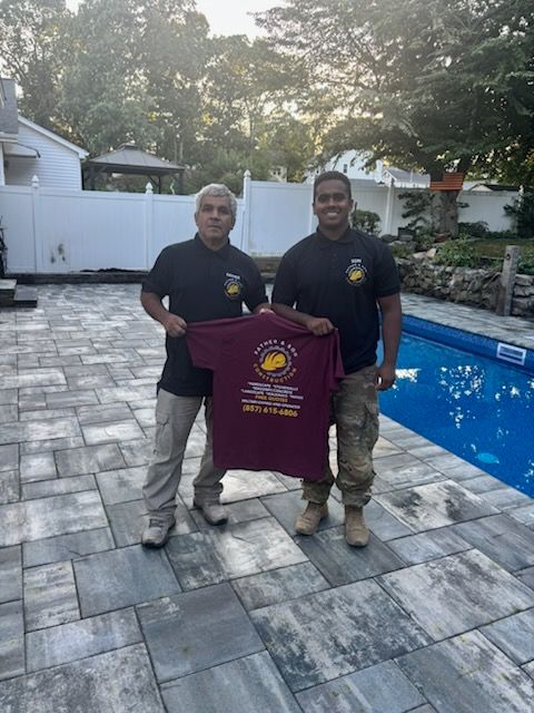 Two men stand by a pool holding a maroon t-shirt. They wear black shirts and khaki pants. Outdoors.