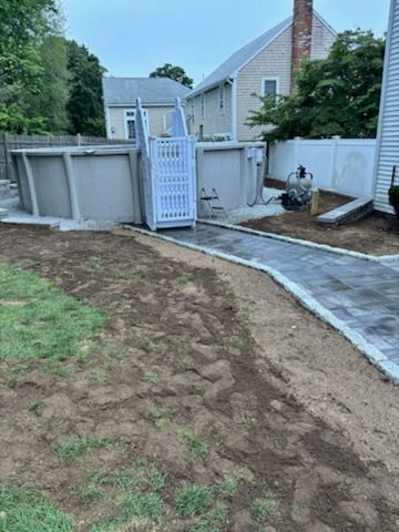 Above-ground pool with a paved walkway, accessible lift, and surrounding grass and houses.