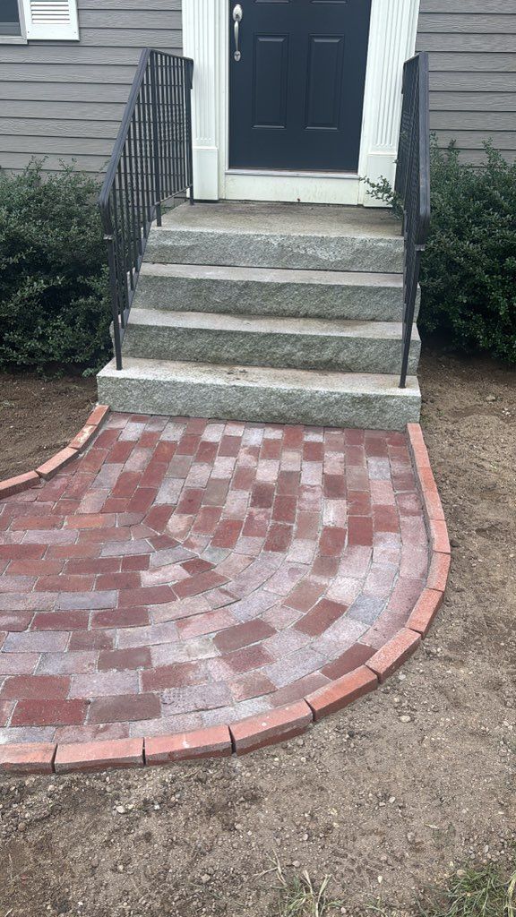Brick path curves to granite steps leading to a dark door with black railing.
