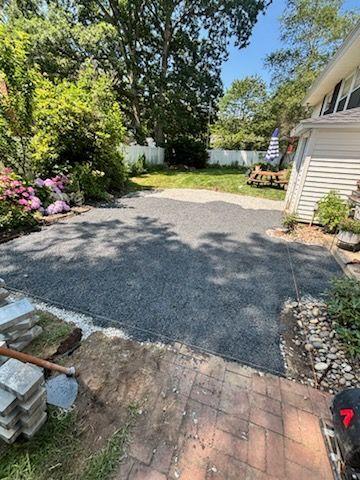 Gravel driveway next to a house, brick patio in foreground, trees and grass in the background.