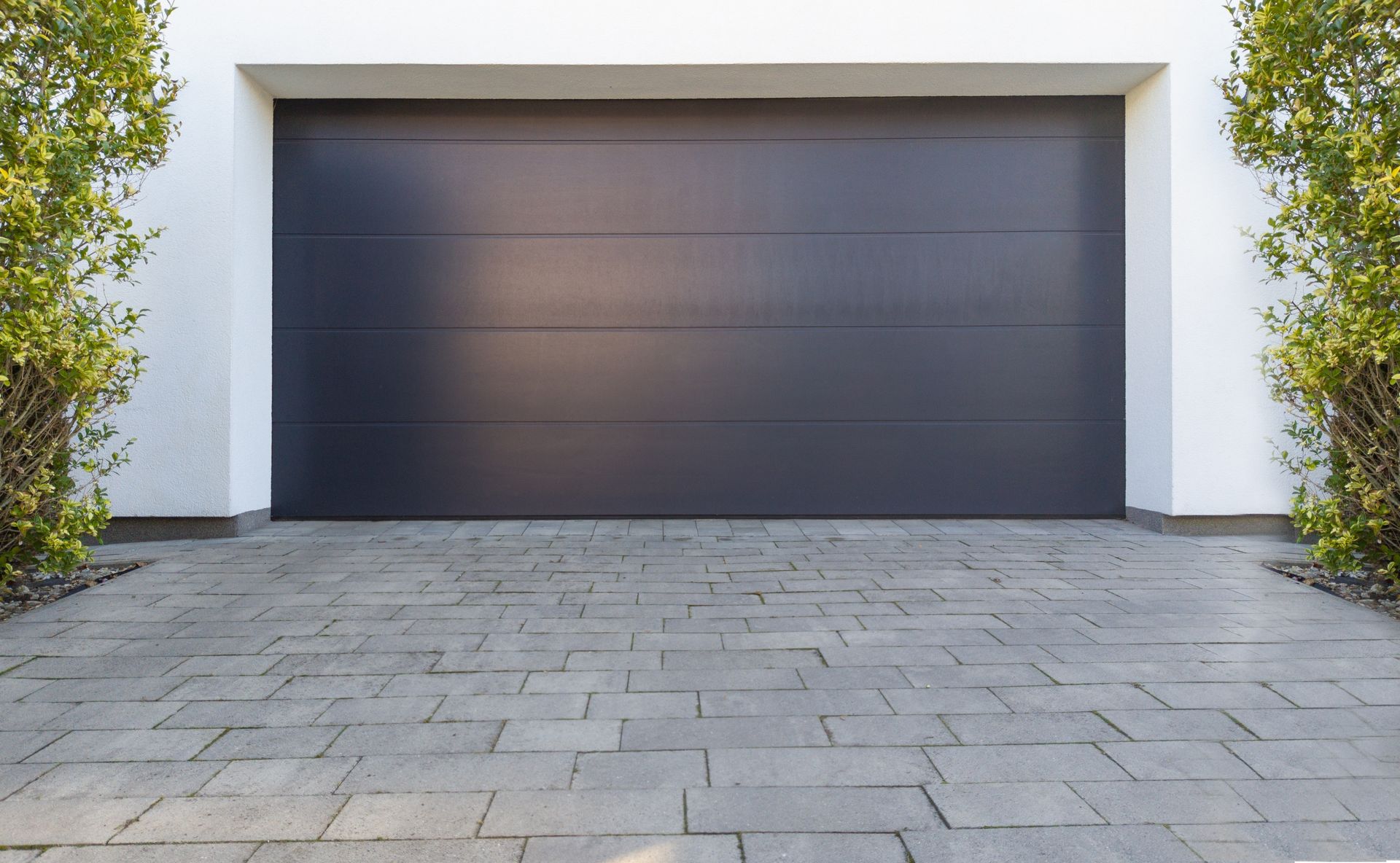 Dark gray garage door with paving stone driveway, white trim, and small green bushes on each side.