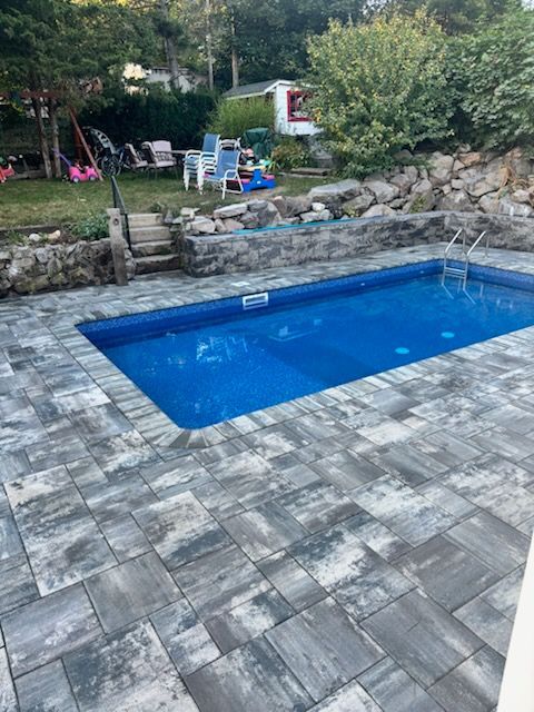 Swimming pool with blue water, surrounded by gray pavers and stone retaining walls; backyard setting.