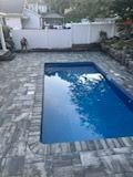 Rectangular blue swimming pool surrounded by gray brick pavers. White fence and house in background.