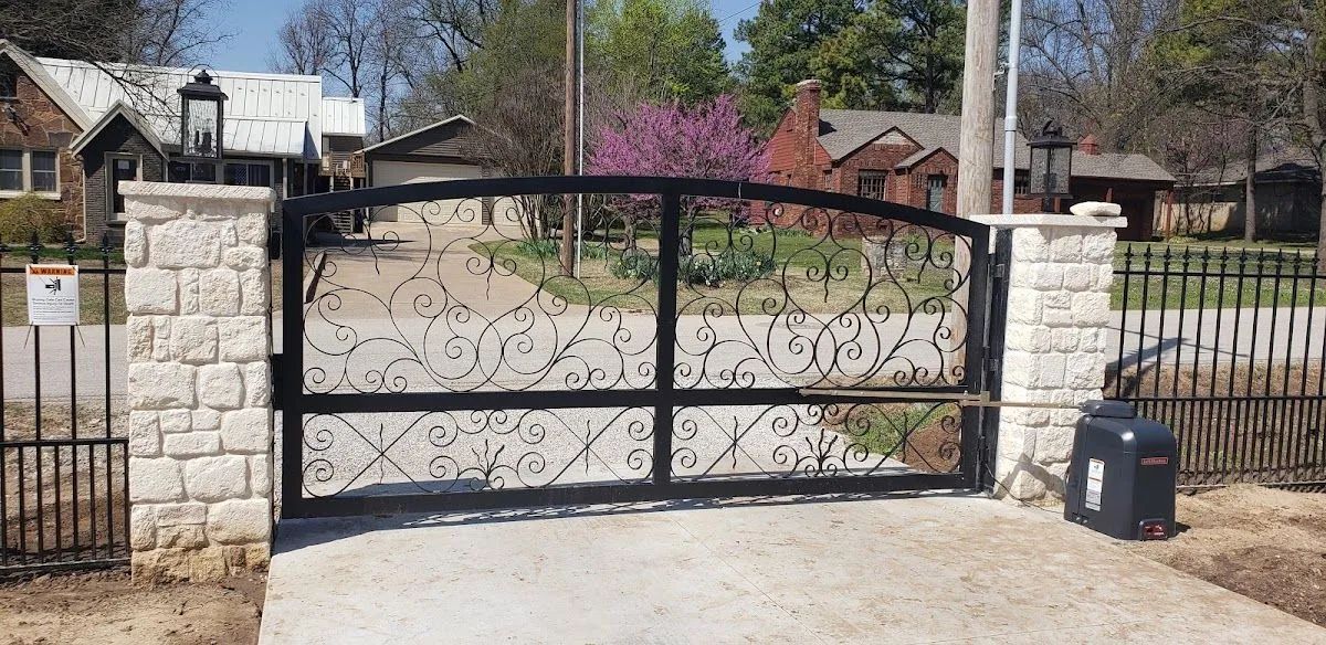 Black decorative wrought-iron gate with stone pillars, leading to a driveway and houses in the background.