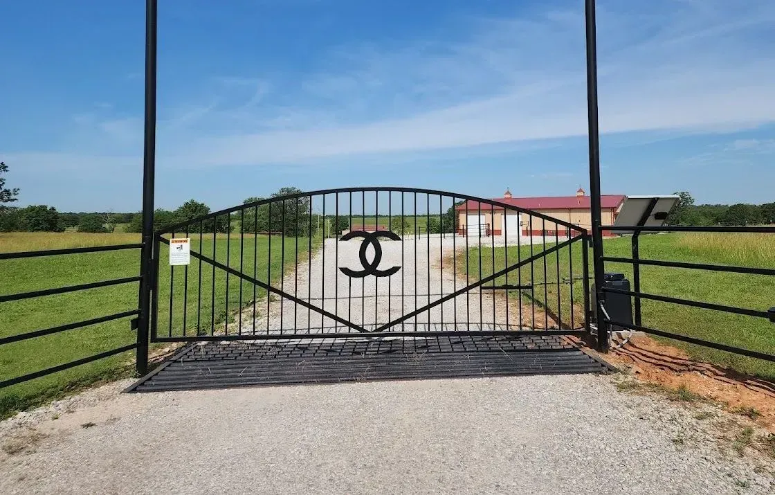 A black gate with a chanel logo on it is open to a dirt road.
