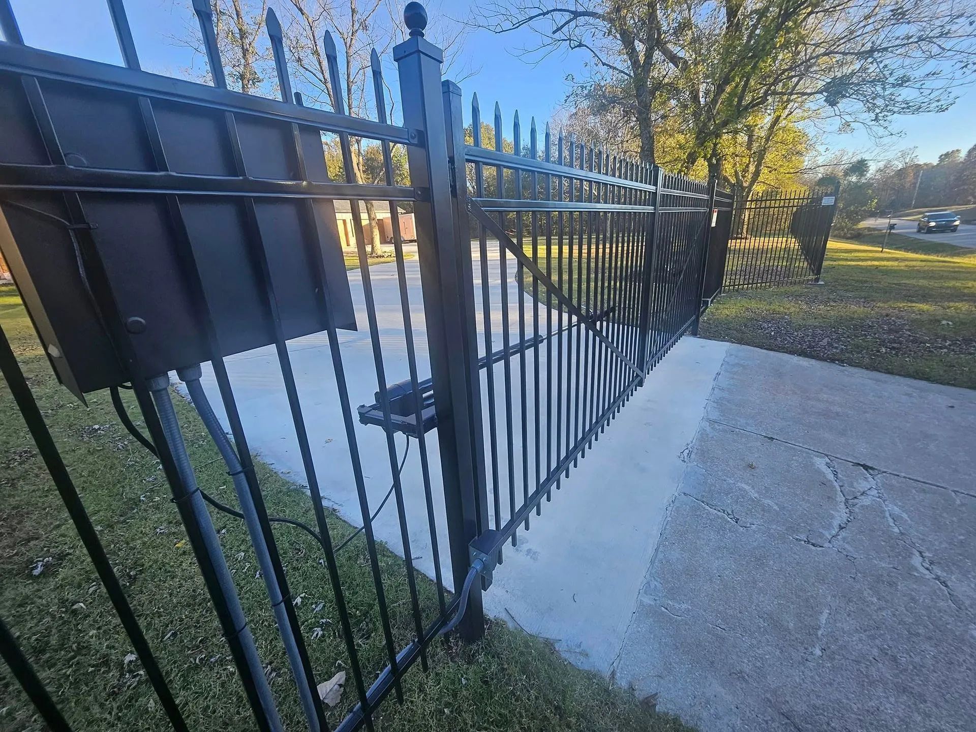 Black metal gate with pointed tops, concrete driveway, and grassy lawn.