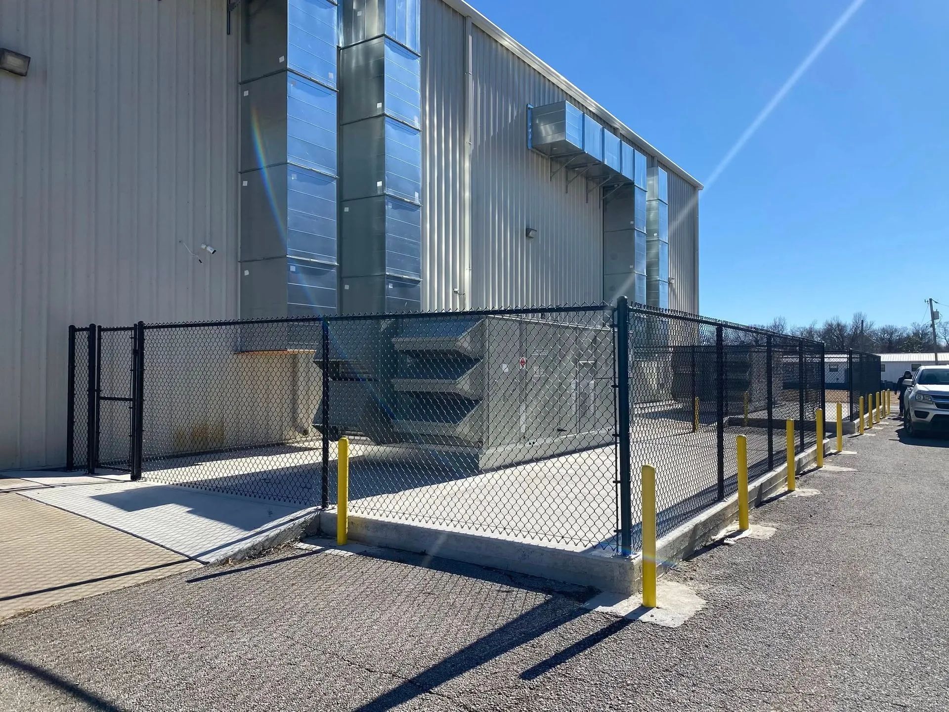 Black chain-link fence surrounds an industrial building with silver ductwork. Yellow bollards line the gravel parking area.