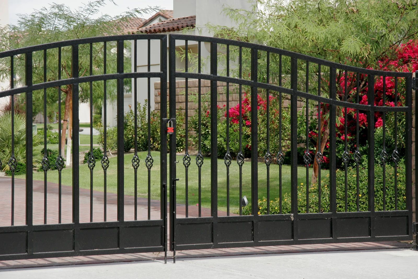A black, arched double-swing metal gate with decorative finials, set in front of a residential home with landscaping.