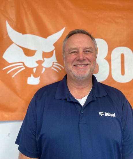 A man in a blue shirt stands in front of a bobcat logo