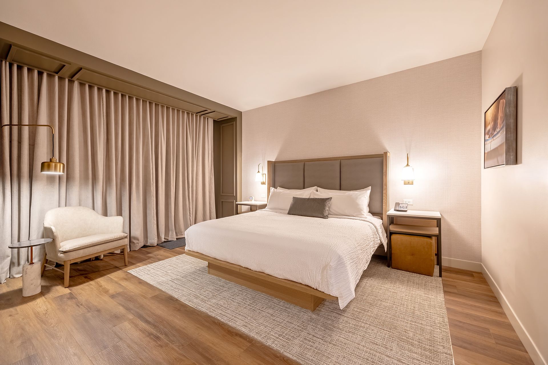 Bedroom with a bed, chair, and floor-to-ceiling curtains; neutral colors, wood flooring.