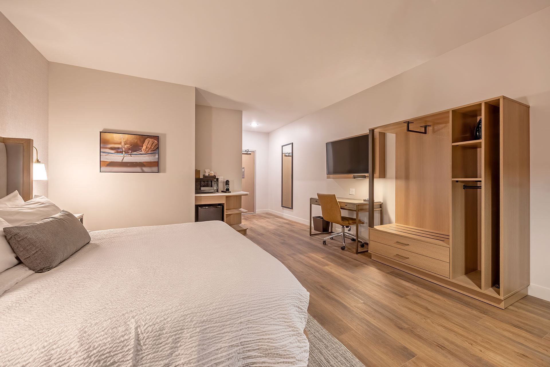 Hotel room with a bed, desk, wardrobe, and a small refrigerator. Neutral color palette and wooden floors.