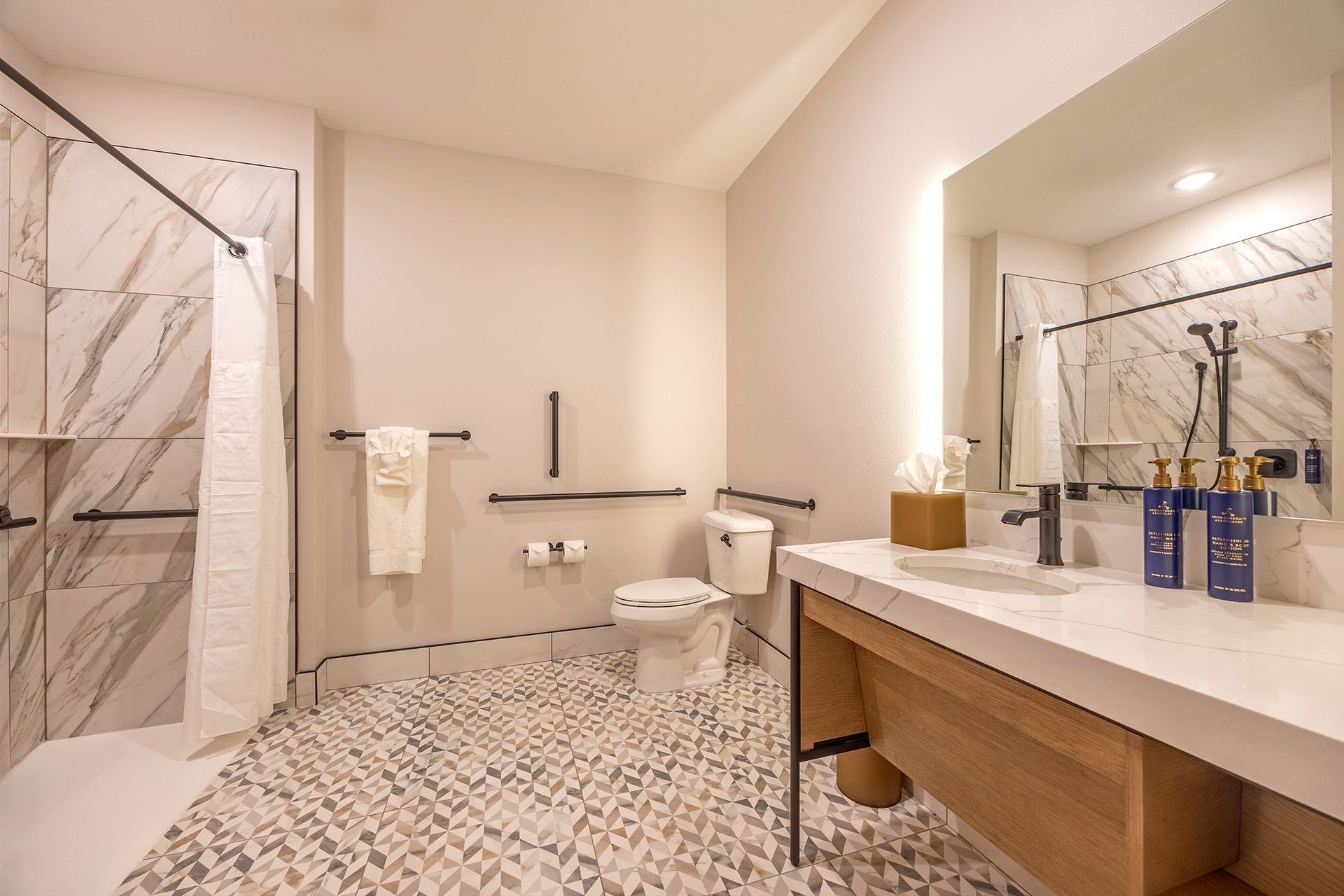 Accessible hotel bathroom with marble shower, grab bars, open vanity, and mosaic tile floor.