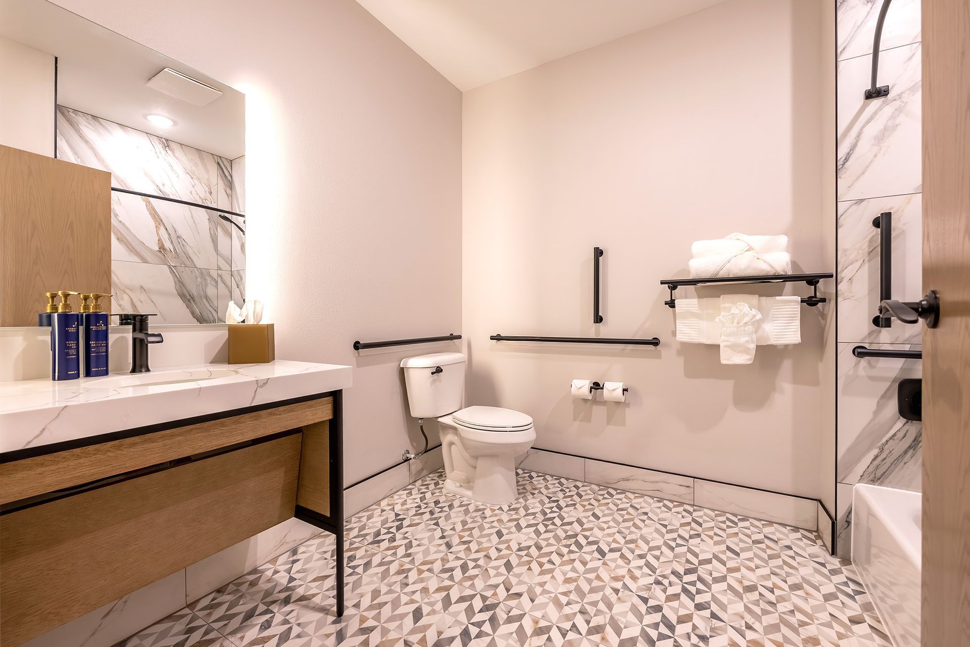 Accessible bathroom with light wood vanity, marble countertop, grab bars, and patterned floor tiles.