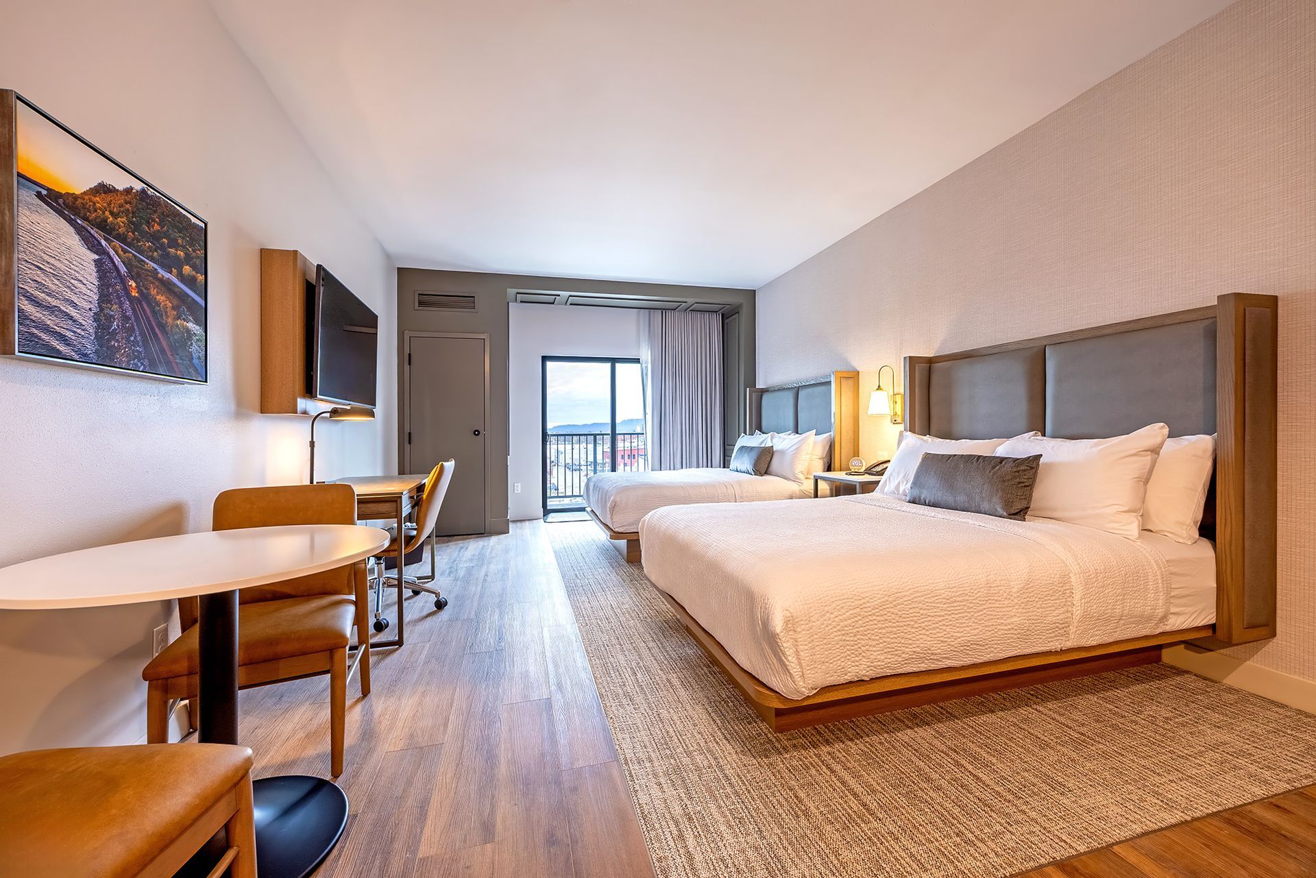 Hotel room with two beds, a desk, table, and balcony overlooking the city.