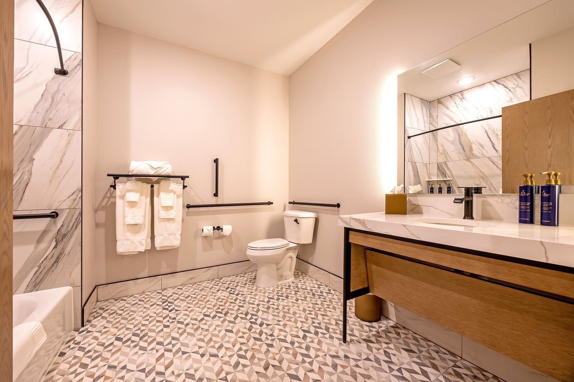 Accessible bathroom with shower, toilet, and sink. White marble walls and countertop. Beige and gray patterned floor.