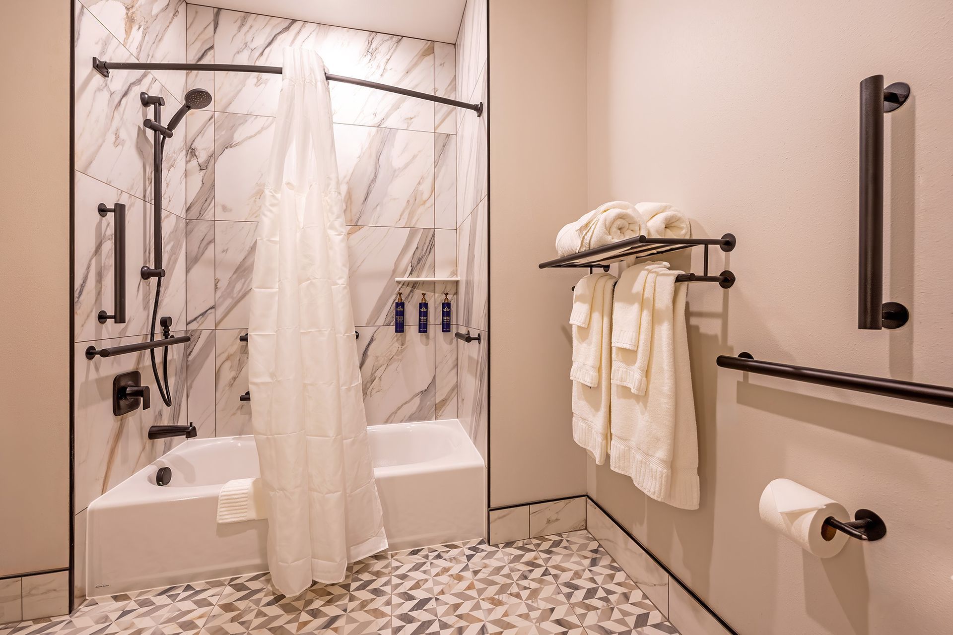 Accessible bathroom with a tub, marble tile, and grab bars. White towels on a rack.