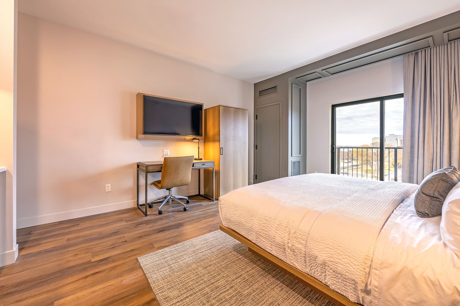Bedroom with bed, desk, TV, closet, and balcony door; light wood floors, rug.