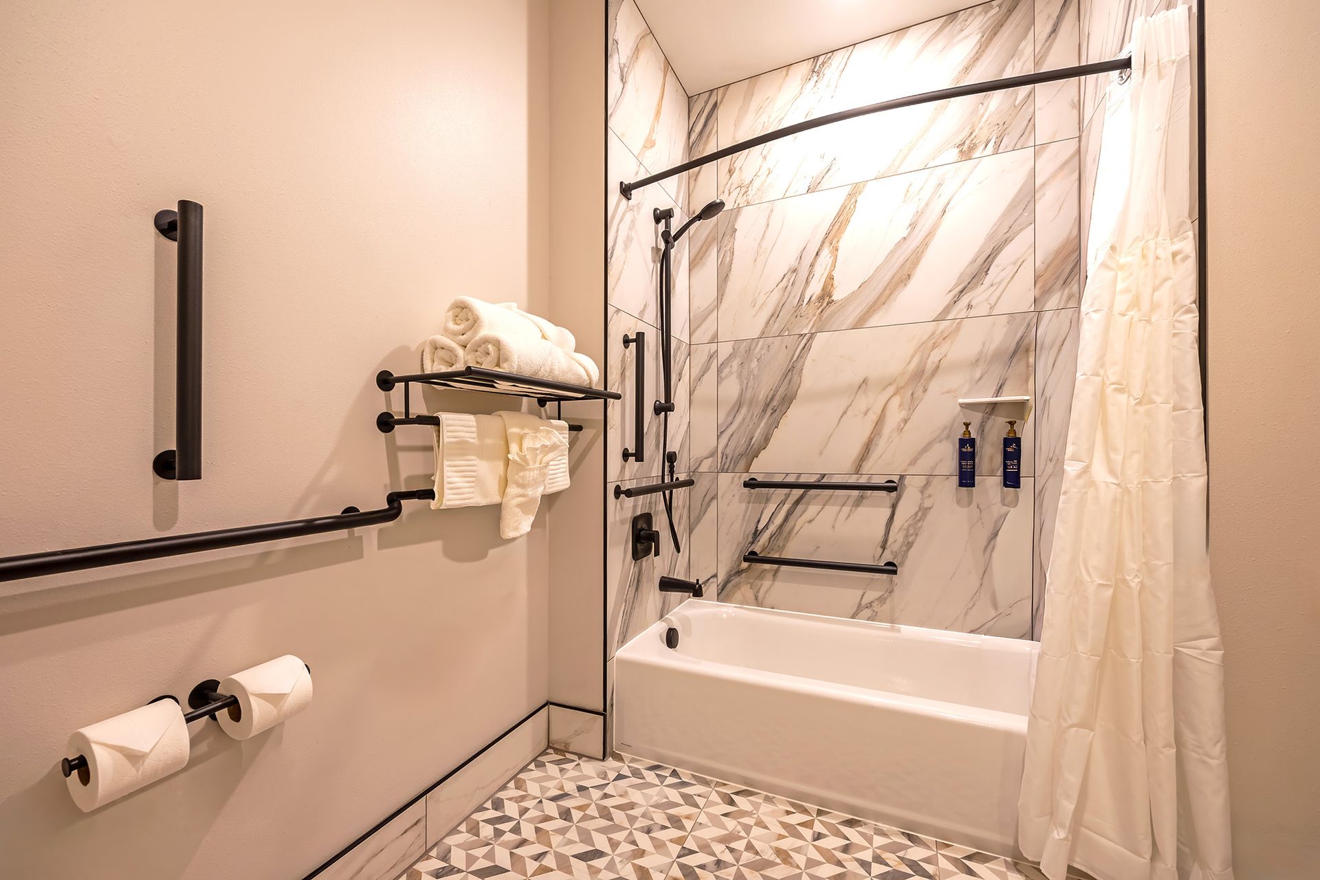 Accessible hotel bathroom with grab bars, shower, and towel rack. Light marble tile and neutral walls.