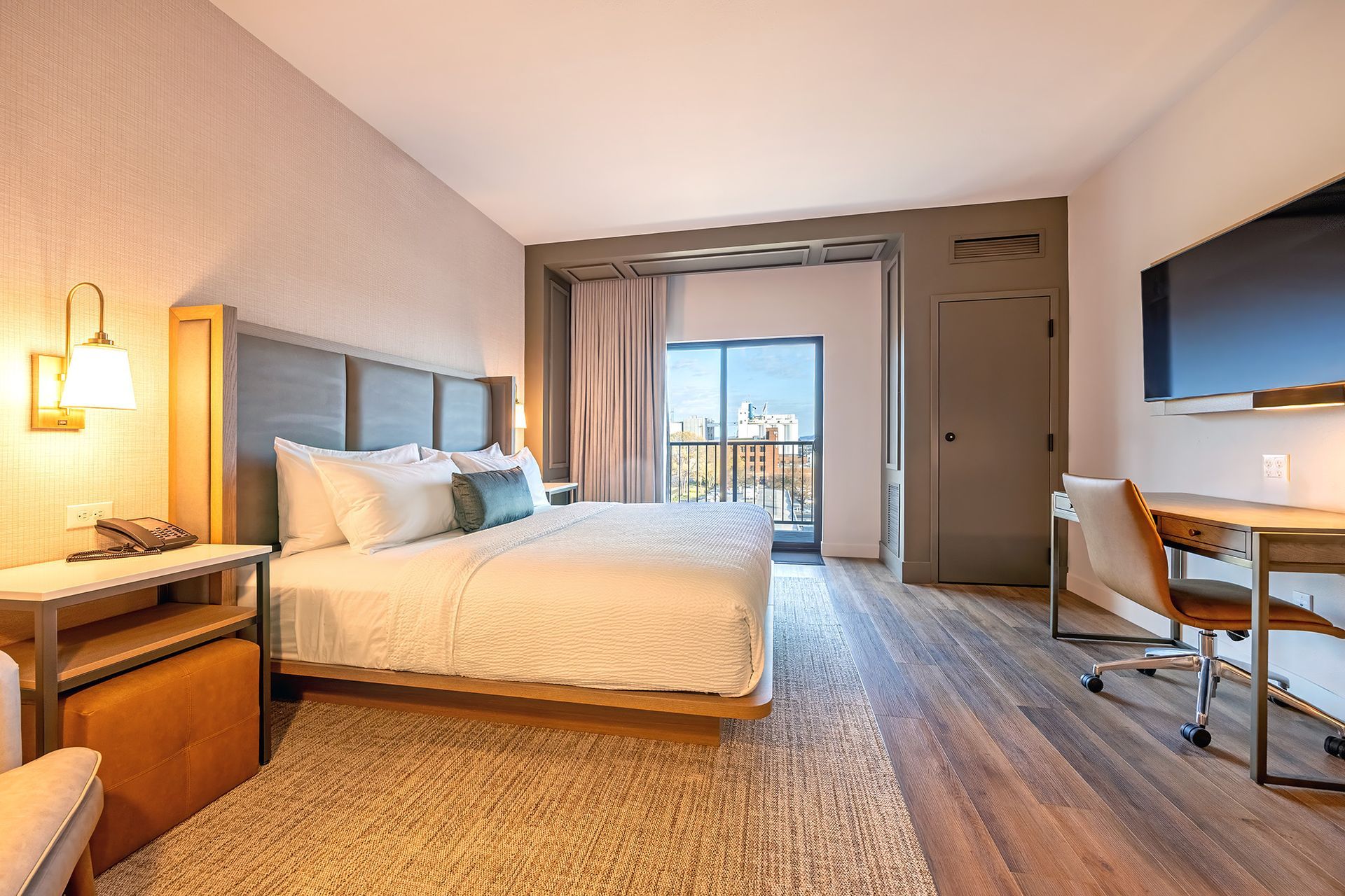Hotel room with a king-sized bed, desk, balcony access, and wood floors.