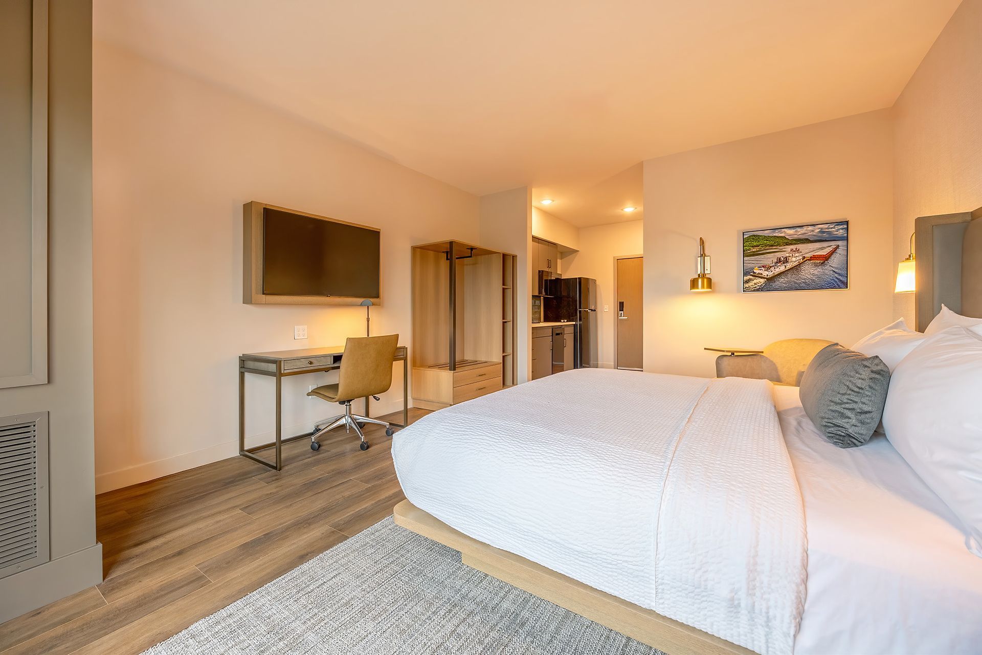 Hotel room with a bed, desk, TV, closet, and kitchenette, soft neutral colors.