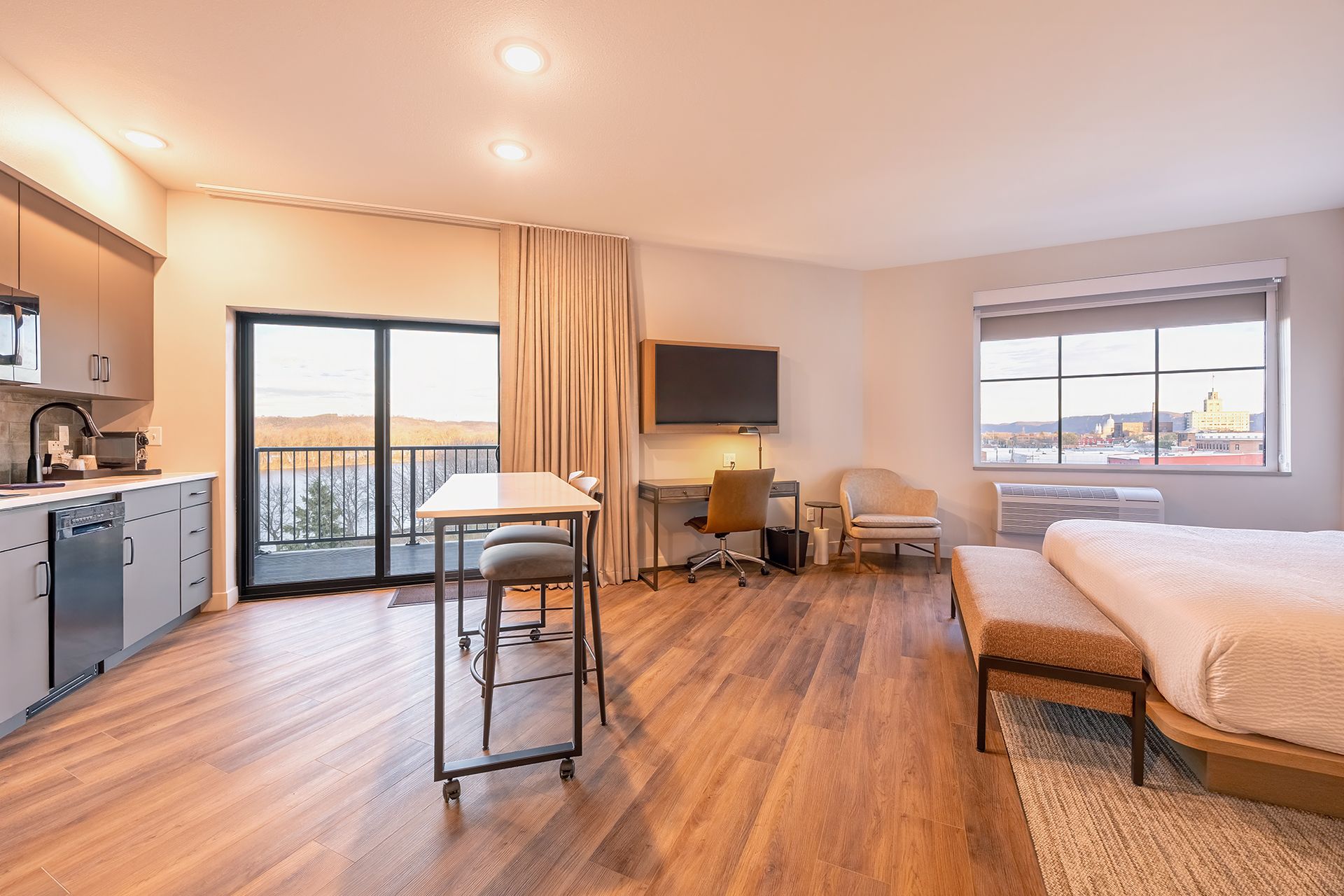 Modern studio apartment with kitchen, desk, bed, and balcony overlooking a scenic river and city view.