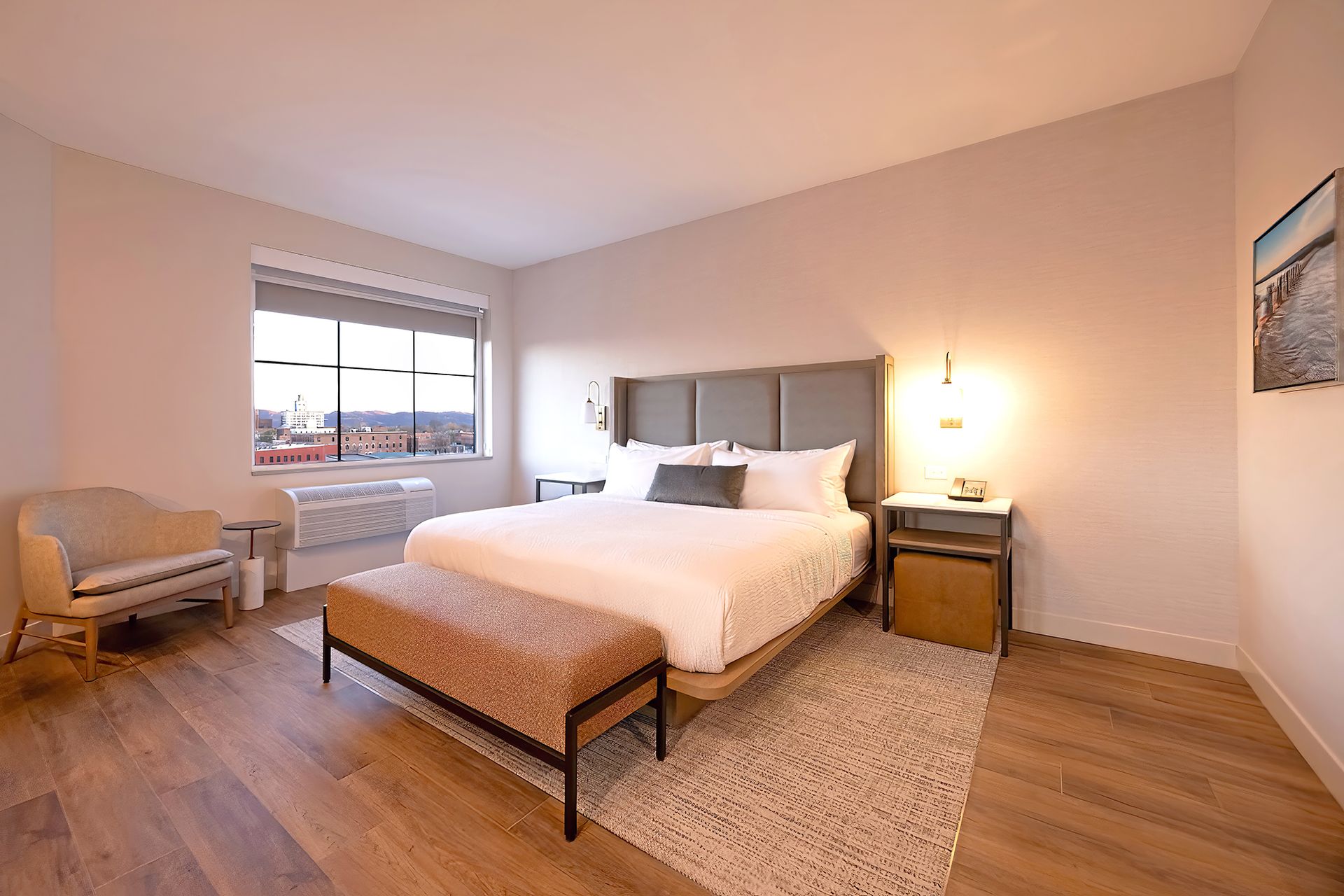 Bedroom with a bed, bench, chair, window, and rug. Warm lighting and wood flooring.