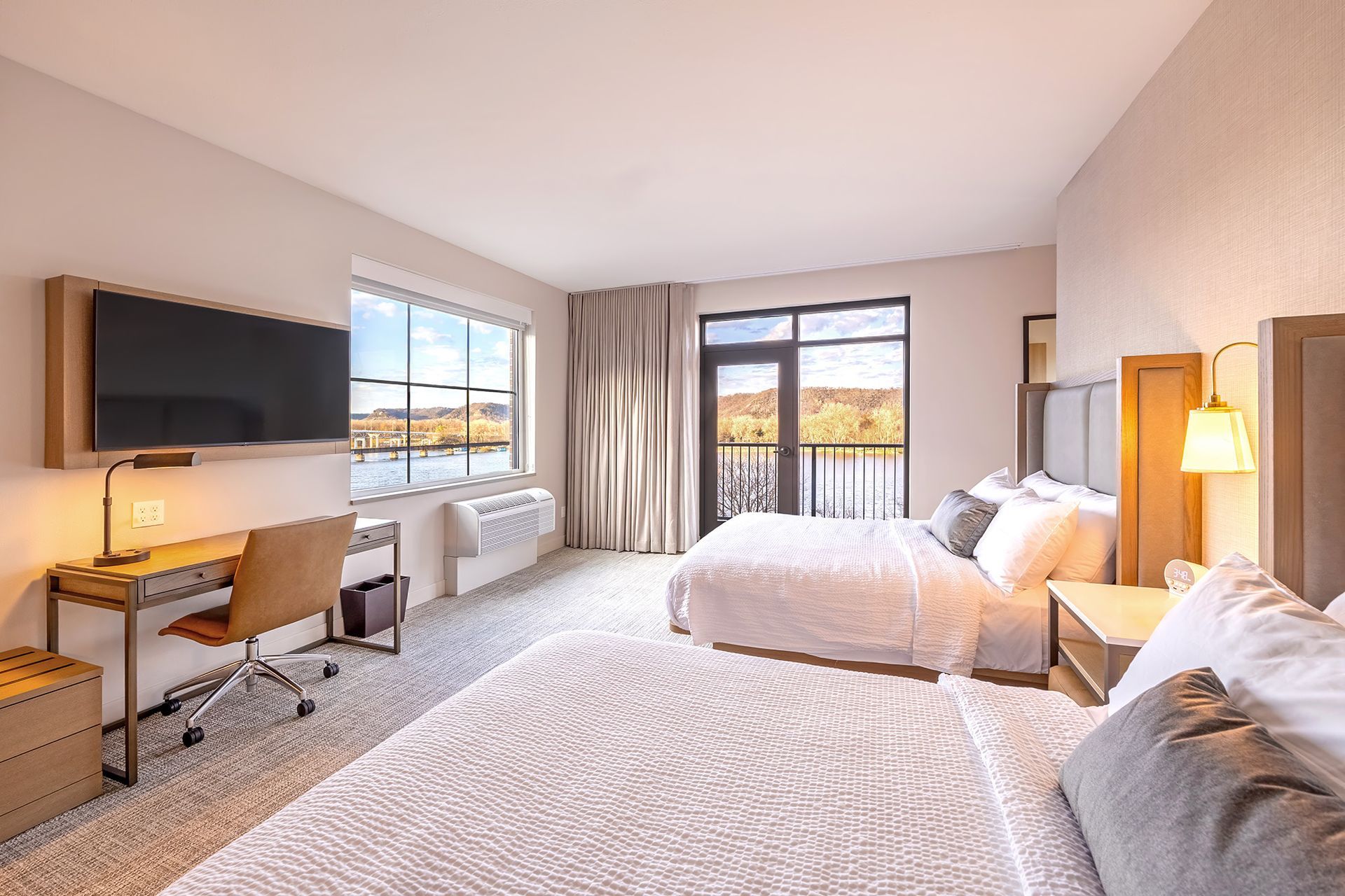 Hotel room with two beds, desk, TV, and a view of the river and bluffs.