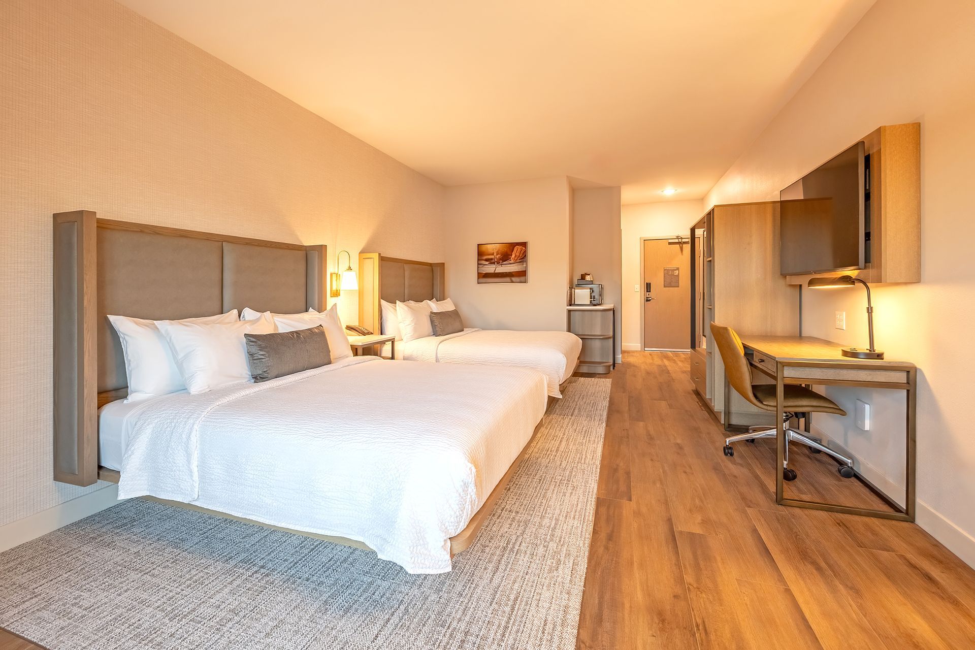 Hotel room with two beds, desk, TV, and wooden floor. Warm lighting.