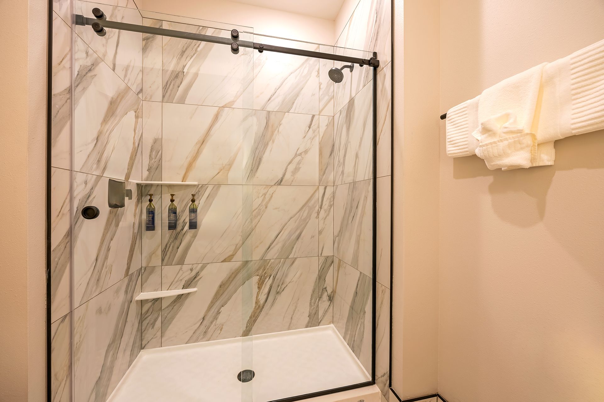 Modern bathroom shower with marble-look tiles, black frame, and glass door. Soap dispensers and shaving shelf.