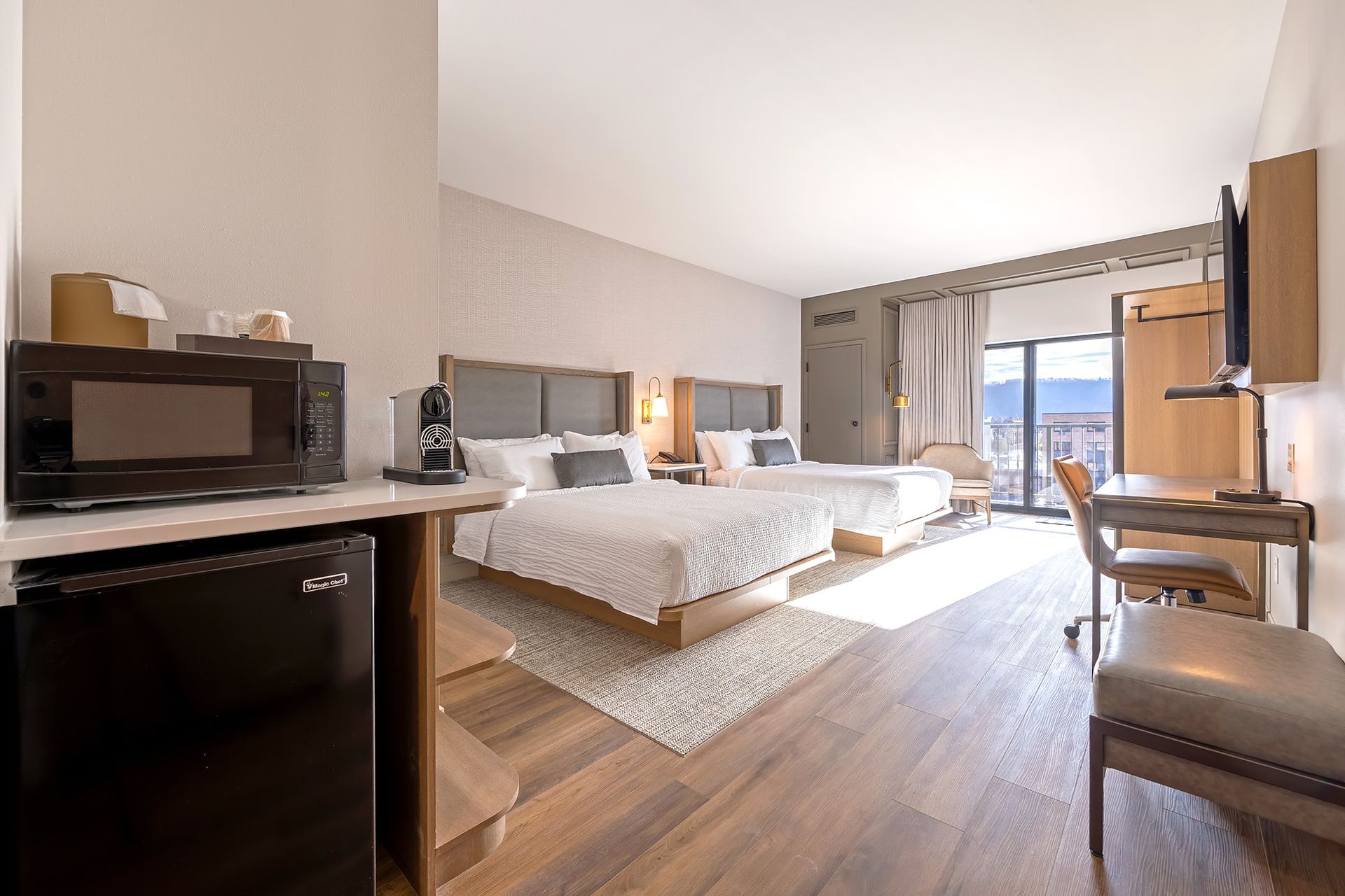 Hotel room with two beds, microwave, mini-fridge, desk, and view of city and bluffs from a balcony.
