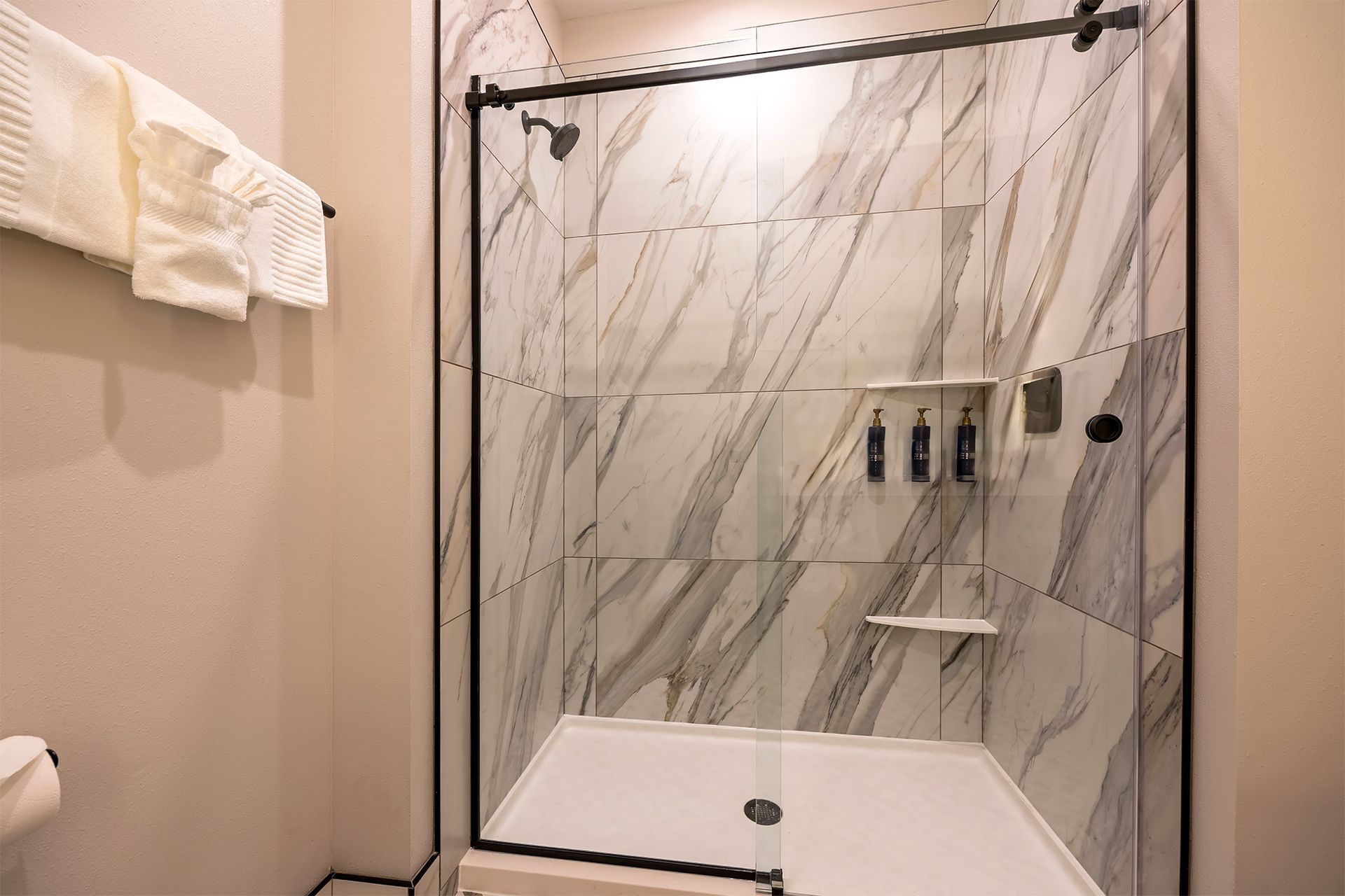 Modern bathroom shower with marble-look tiles, black frame, and glass door. Soap dispensers and shaving shelf.