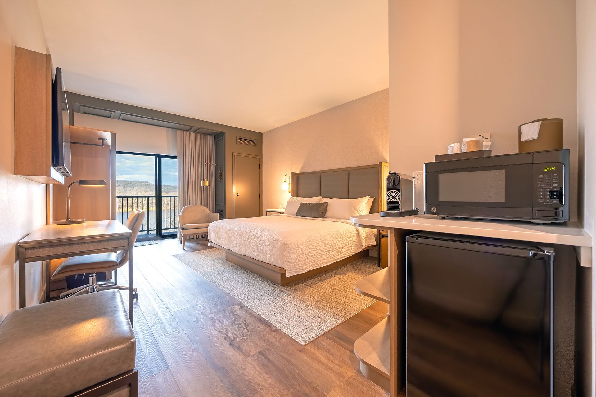 Hotel room with a king bed, desk, microwave, mini-fridge, and balcony overlooking a scenic view.