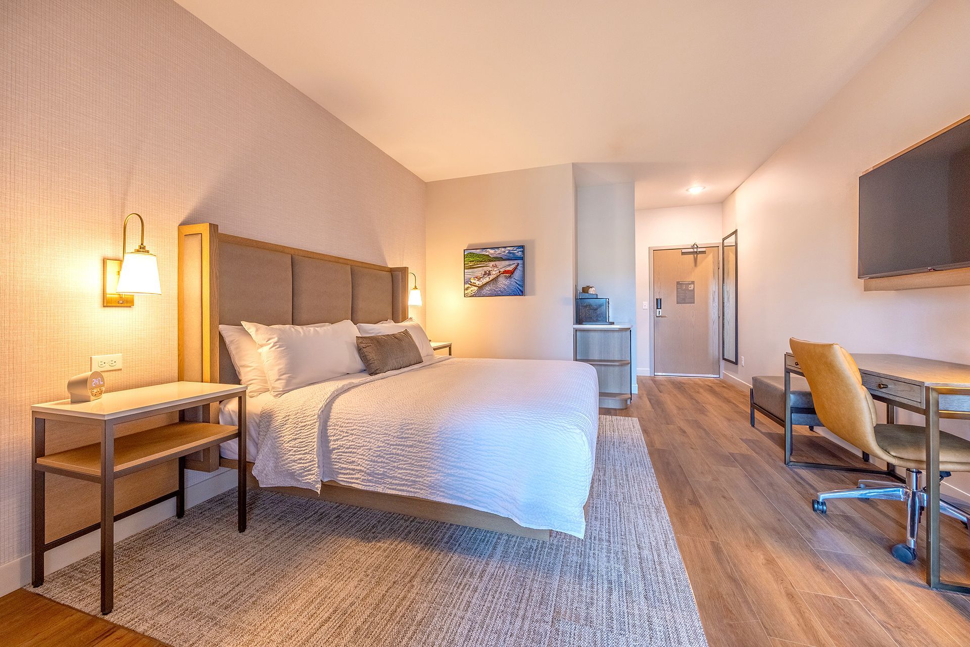 Hotel room with a bed, desk, and side table. Neutral colors and wood flooring.
