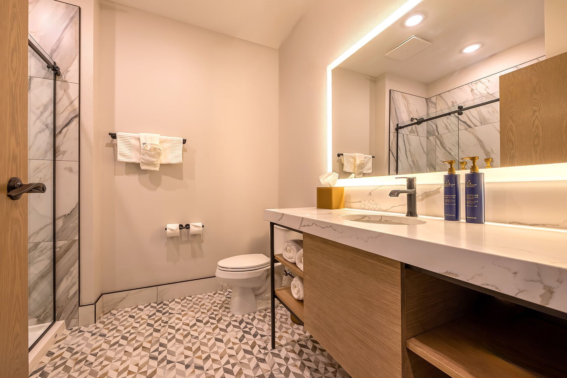 Hotel bathroom with marble shower and countertop, wooden vanity, and patterned tile floor.