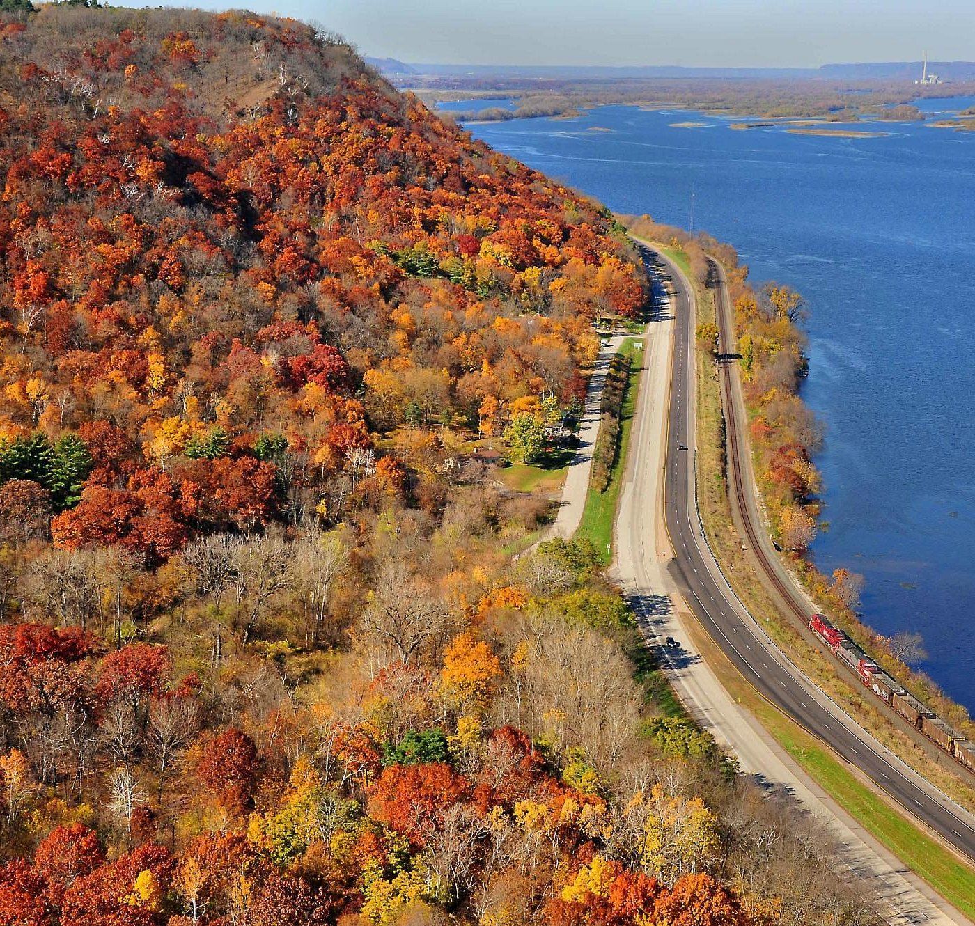 Autumn foliage along a road next to a river. Red, orange and yellow leaves cover the hillside.