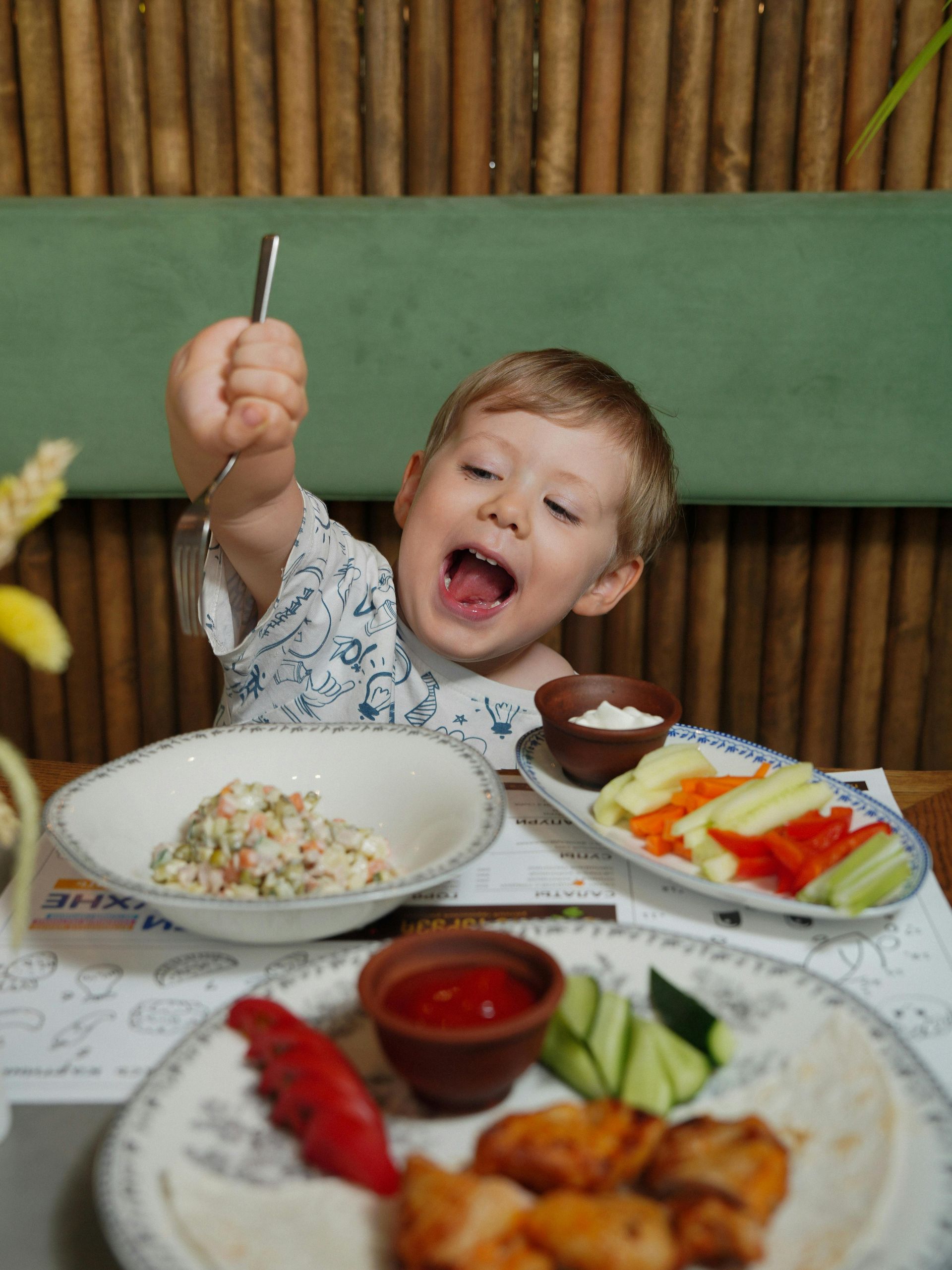 A toddler with an excited expression reaches up with a spoon while sitting at a table with several plates of food.