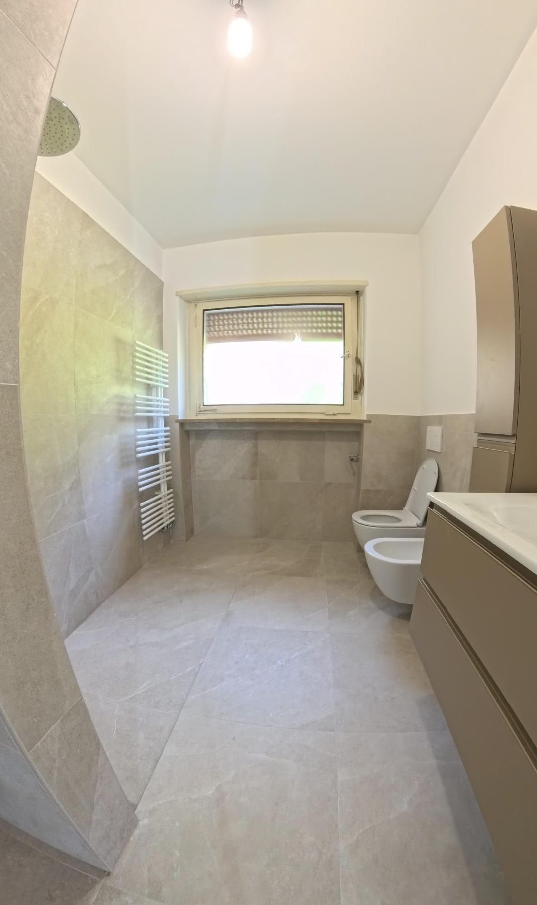 Bagno moderno con piastrelle beige, doccia, WC, bidet, mobile lavabo e finestra.