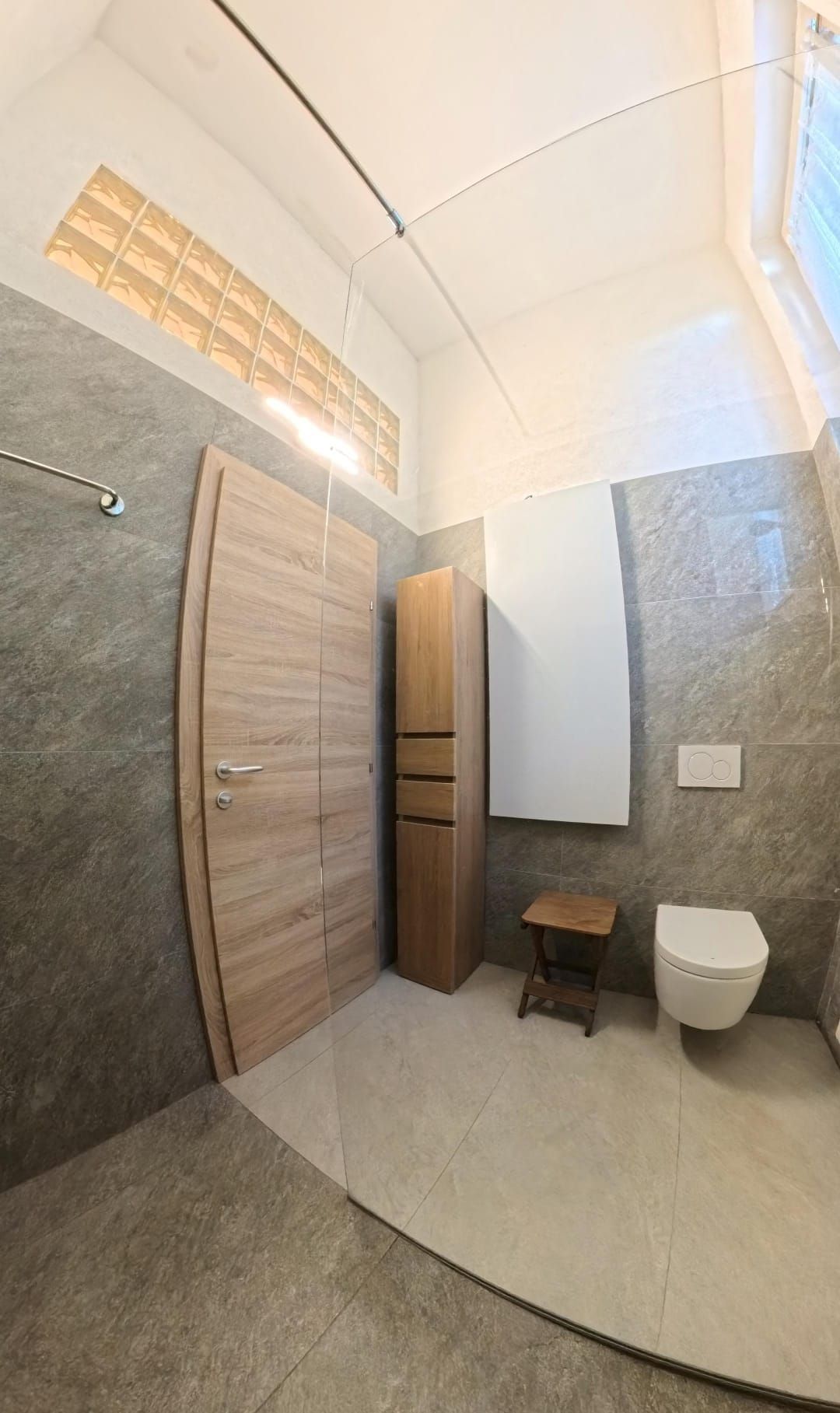 Bagno con pareti piastrellate grigie, porta e mobiletto in legno chiaro e WC bianco.
